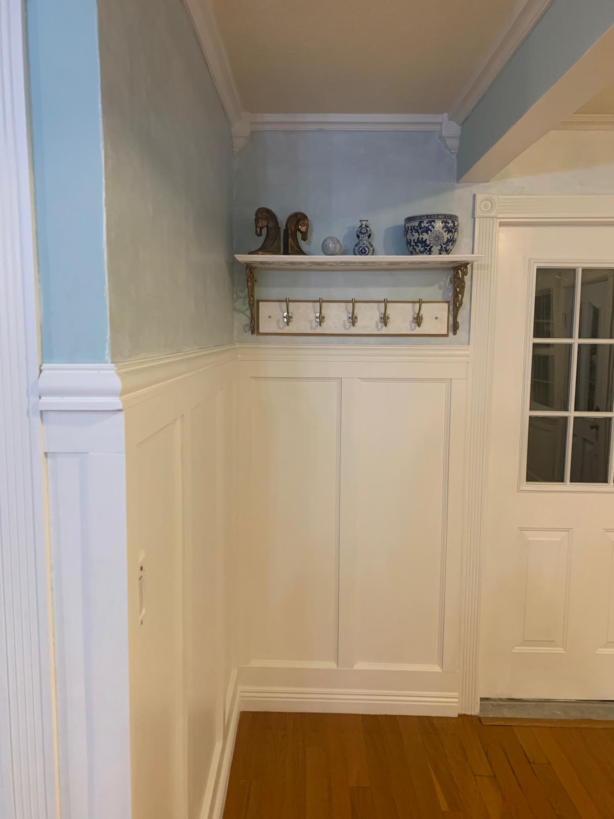 Finished farmhouse entry corner with paneled walls, brass hooks, a decorative shelf, and blue-and-white ceramics